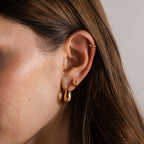 Close-up of a woman's ear wearing three gold earrings: a hoop, a small stud, and the Love Knot Huggies—teardrop-shaped earrings perfect for celebrating lasting bonds or giving as a meaningful gift.