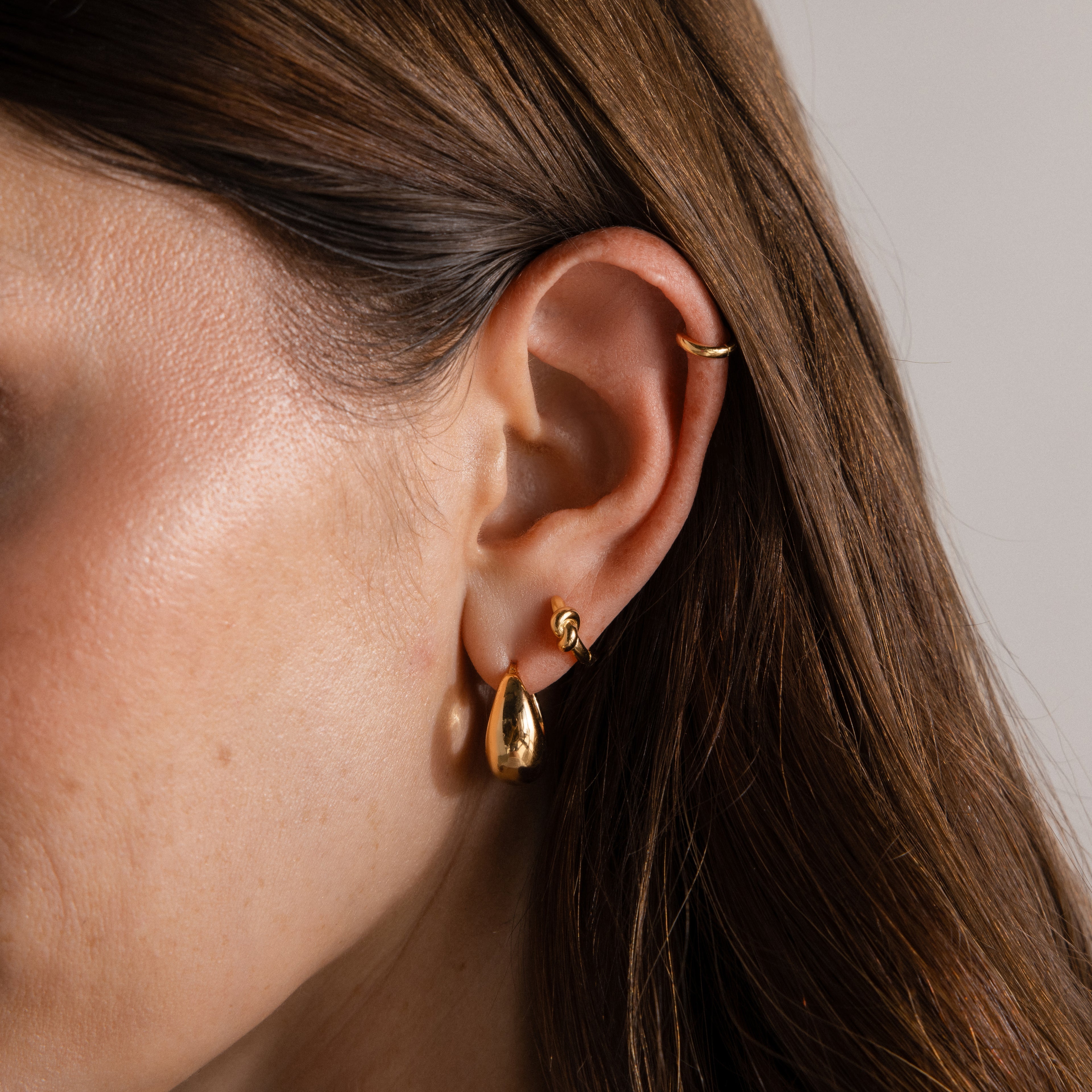Close-up of a woman's ear wearing three gold earrings: a hoop, a small stud, and the Love Knot Huggies—teardrop-shaped earrings perfect for celebrating lasting bonds or giving as a meaningful gift.