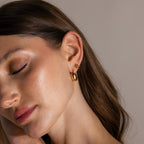 Woman with closed eyes wears the Love Knot Huggies in 18K Gold and an ear cuff, touching her face against a neutral background—a meaningful gift that celebrates lasting bonds.