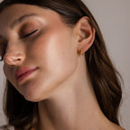 With eyes closed and head tilted, a woman wears the Love Knot Huggies in 18K Gold—an elegant earring and subtle symbol of lasting bonds, making a meaningful gift.