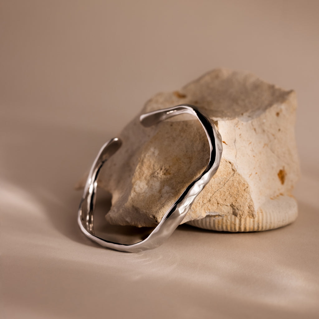 The Organic Wavy Cuff Bracelet in Silver, with sculpted curves and a textured finish, rests on a light-colored, textured rock against a neutral background.