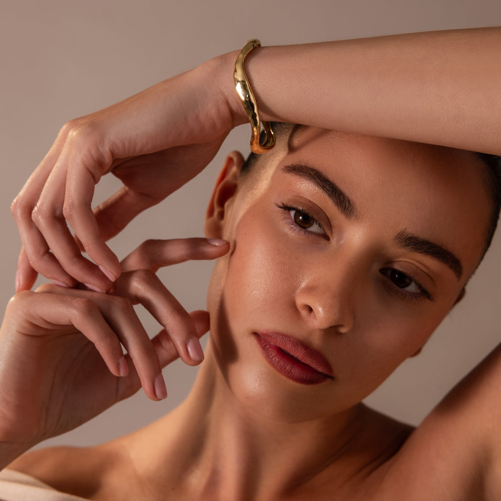A woman with natural makeup poses with hand on forehead, wearing the Organic Wavy Cuff Bracelet in 18K Gold—a timeless accessory that highlights her sculpted curves as she gazes thoughtfully aside.
