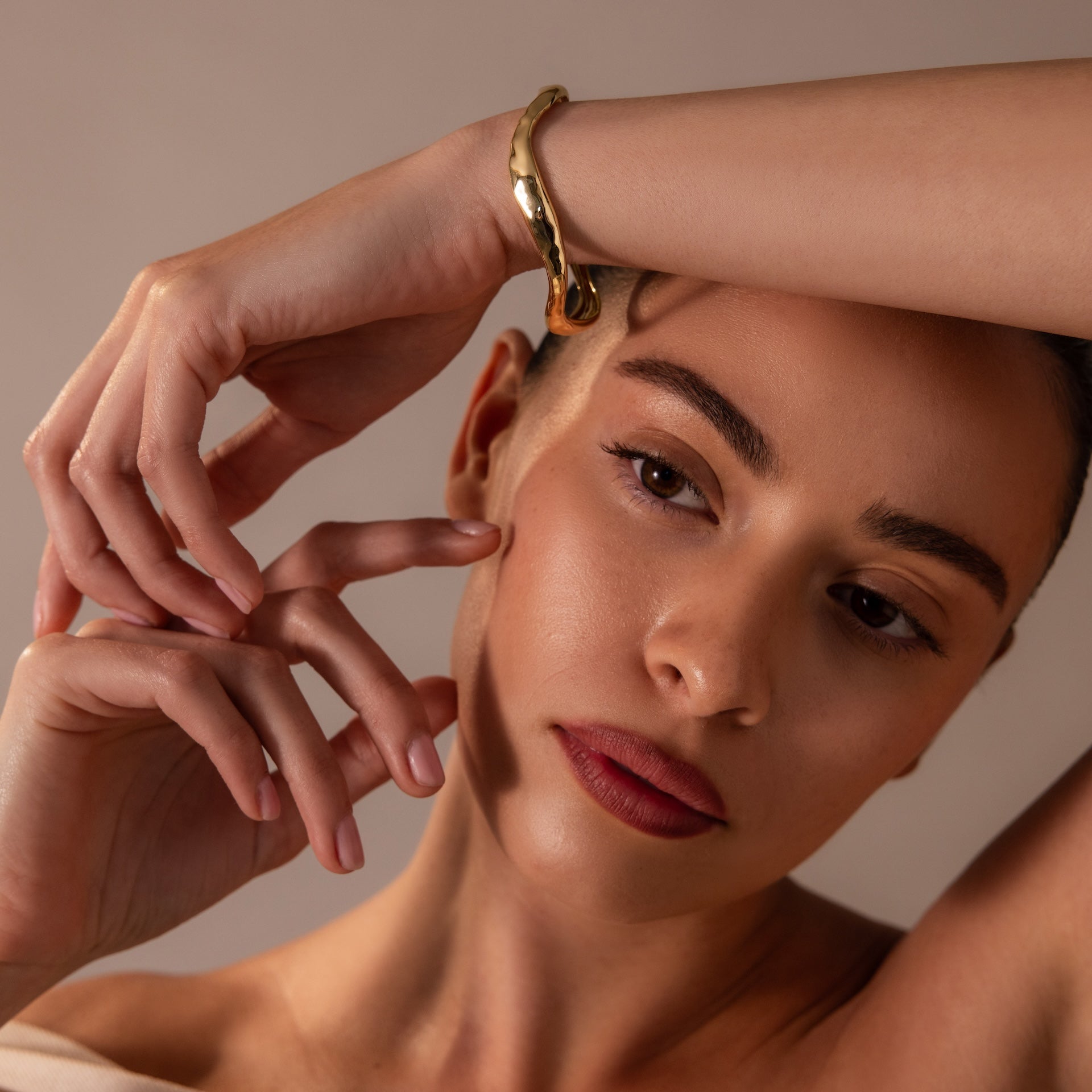 A woman with natural makeup poses with hand on forehead, wearing the Organic Wavy Cuff Bracelet in 18K Gold—a timeless accessory that highlights her sculpted curves as she gazes thoughtfully aside.