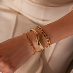A hand wearing the Organic Mariner Chain Bracelet along with two gold bracelets, all featuring unique textures and designs, stands out against a cream outfit—showcasing a modern jewelry aesthetic.