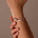 A hand wears the Organic Wavy Cuff Bracelet in Silver, its sculpted curves displayed as another hand gently touches the wrist, all set against a neutral background.