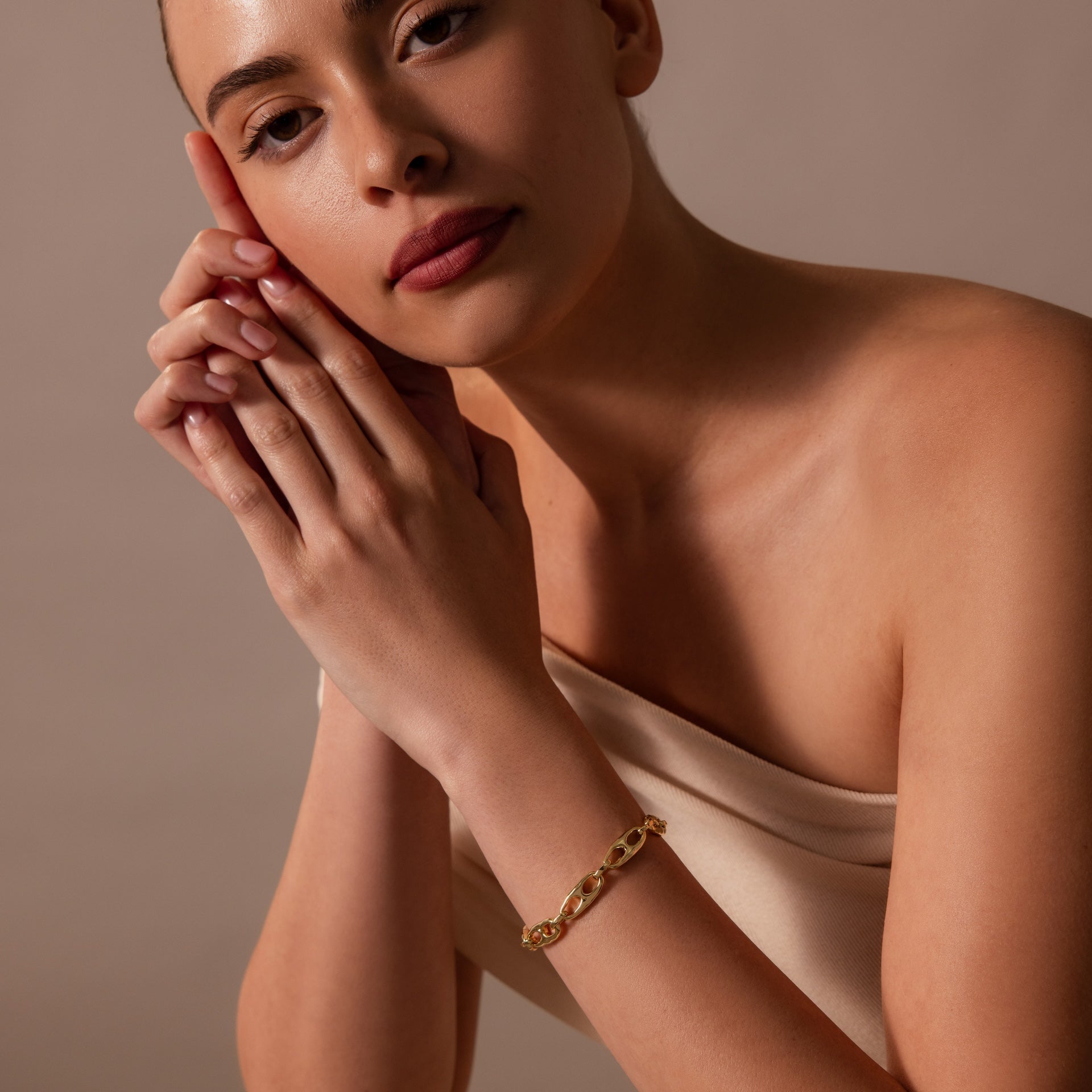 A woman in an off-shoulder top poses with hands clasped, highlighting the Organic Mariner Chain Bracelet in 18K Gold—a sculptural essential—against a neutral background.
