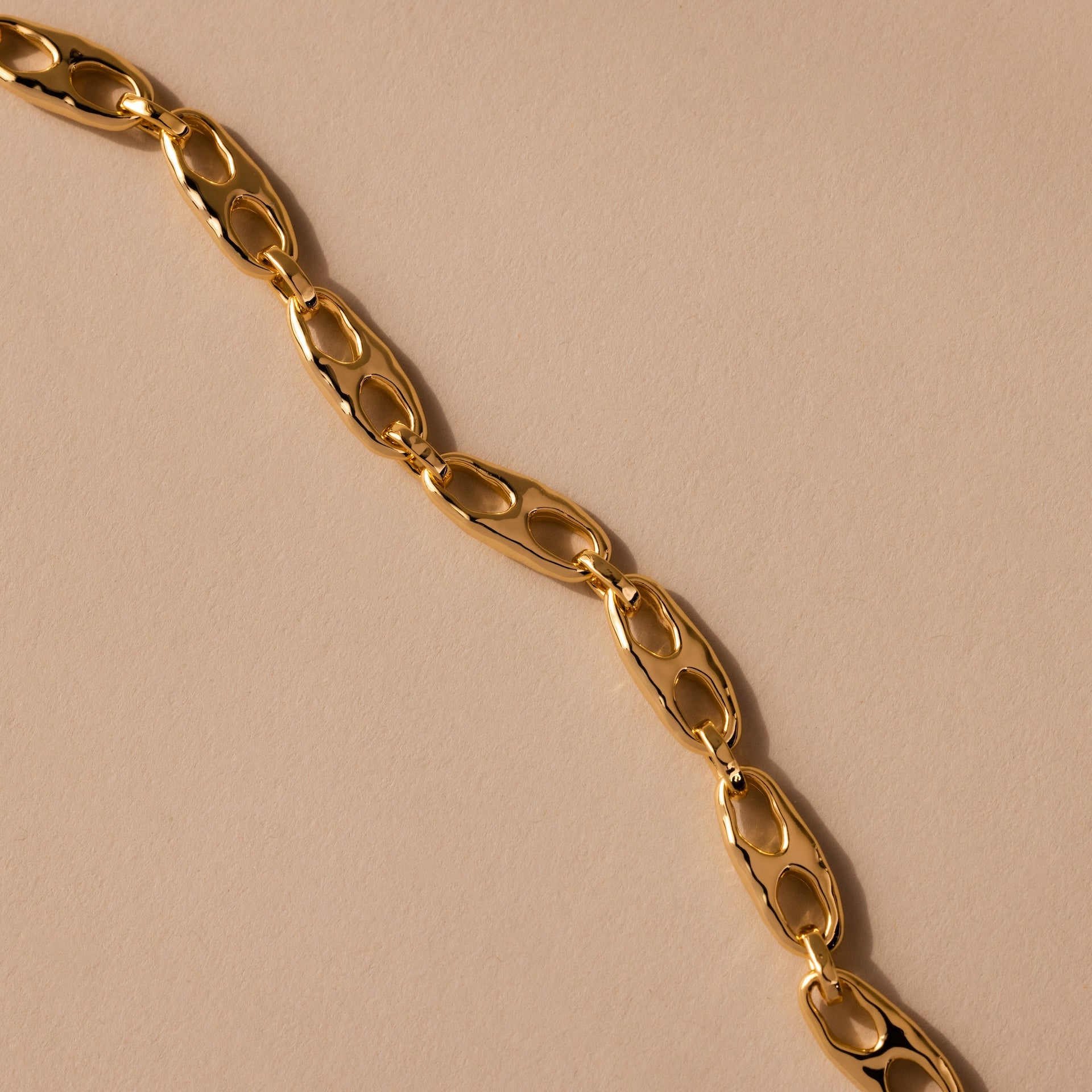 The Organic Mariner Chain Bracelet in 18K Gold features sculptural oval links arranged diagonally, creating a timeless and elegant design against a beige background.