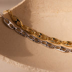 The Organic Mariner Chain Bracelet, available in gold and silver tones, is showcased on a textured beige surface—ideal for fans of contemporary jewelry.
