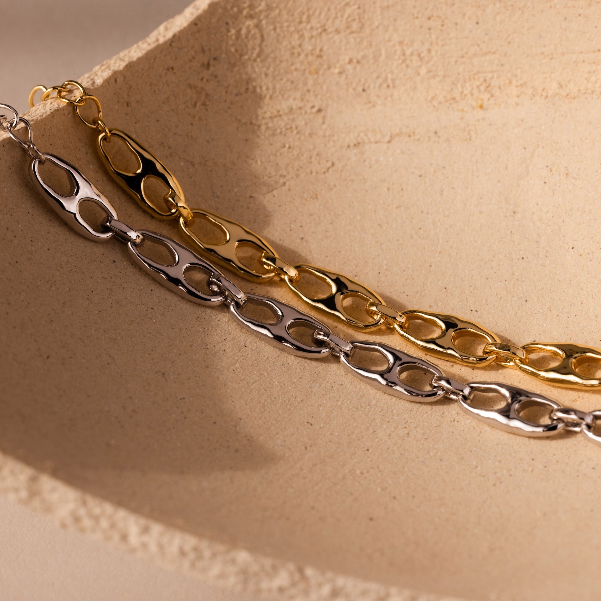 The Organic Mariner Chain Bracelet, available in gold and silver tones, is showcased on a textured beige surface—ideal for fans of contemporary jewelry.