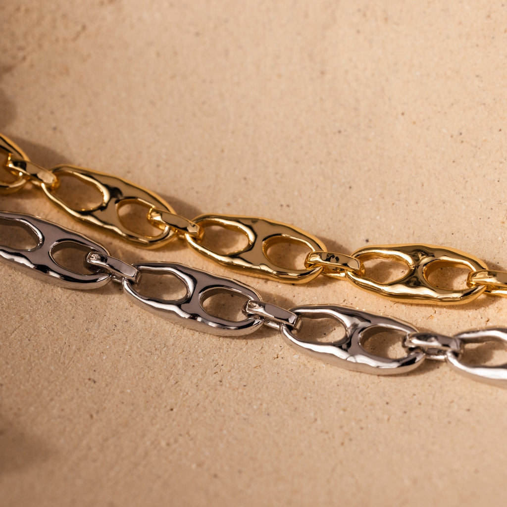 The Organic Mariner Chain Bracelet in 18K Gold and a silver sculptural chain bracelet are displayed on a beige textured surface, highlighting modern jewelry design.