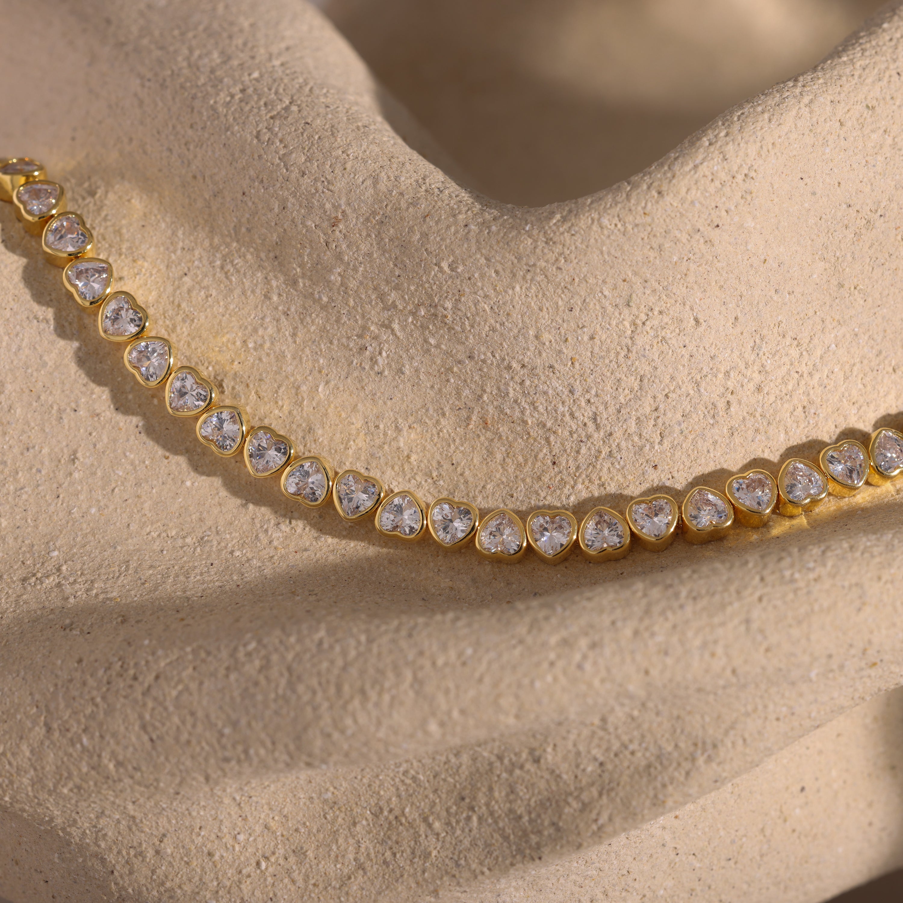 Elegant jewelry showcasing the Heart Tennis Bracelet in 18K Gold, adorned with heart-shaped diamonds and displayed on a textured beige stone surface.
