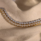 A gold and silver diamond necklace rests on a textured beige surface, elegantly paired with the Heart Tennis Bracelet—a sophisticated accessory featuring heart-shaped stones perfect for complementing any jewelry collection.
