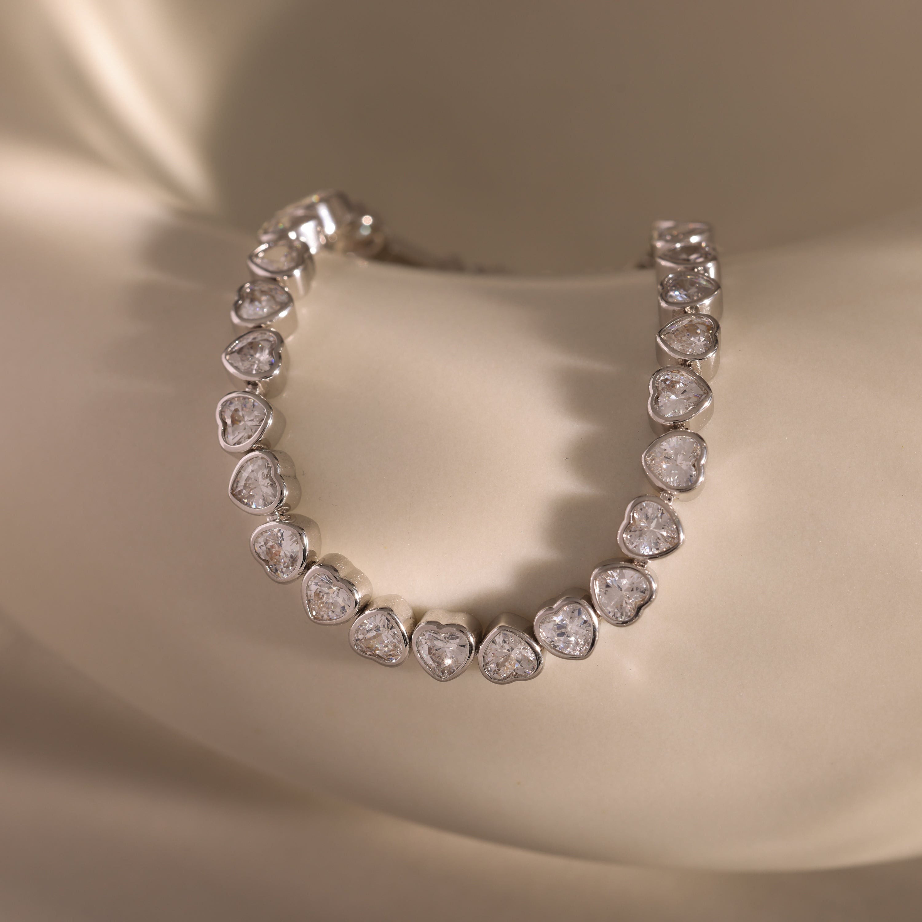 The Heart Tennis Bracelet features heart-shaped stones and sparkling diamonds, elegantly displayed on a smooth, cream-colored surface—making it a stunning and refined jewelry piece.