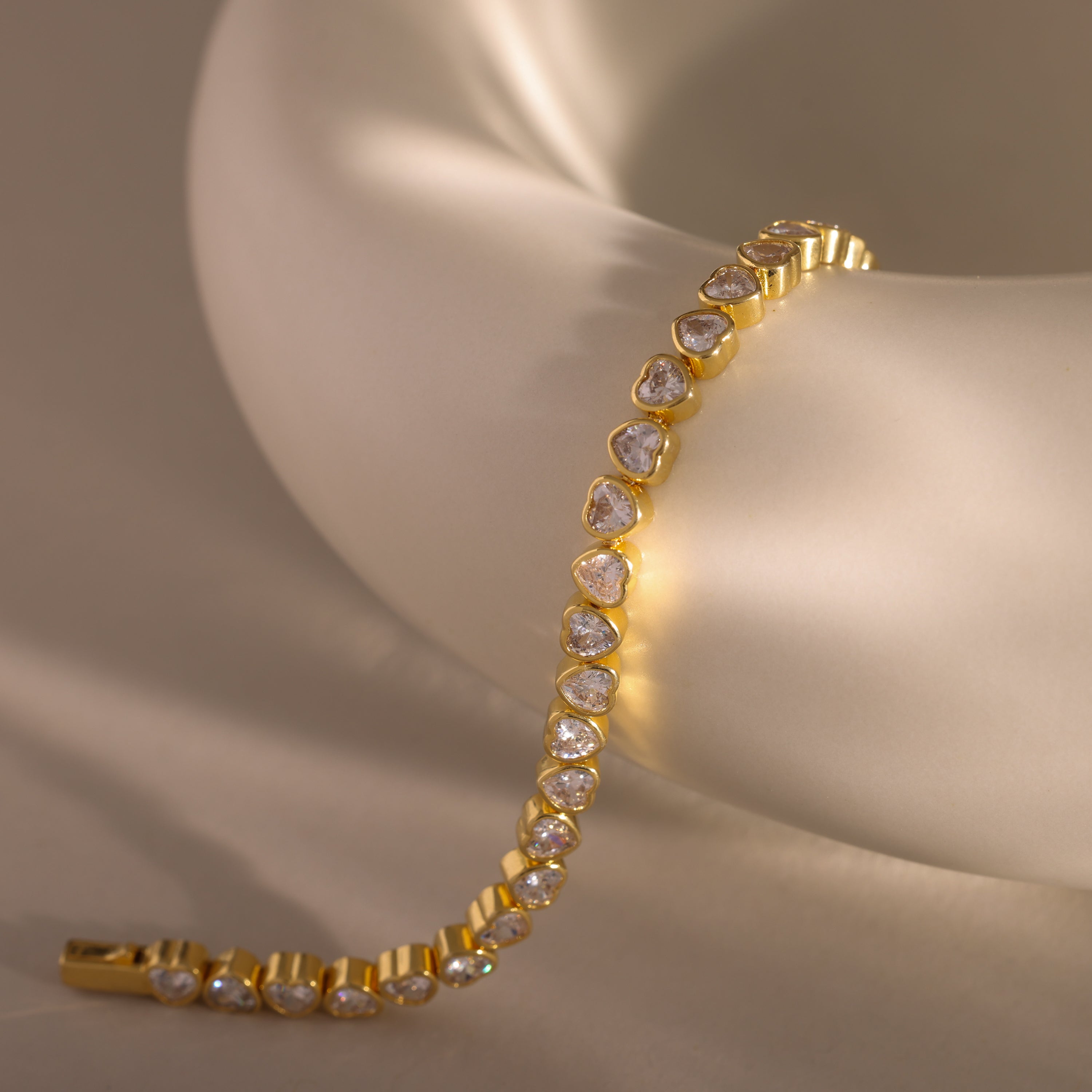 The Heart Tennis Bracelet, an elegant gold piece adorned with sparkling stones, is gracefully displayed on a smooth white ceramic surface.