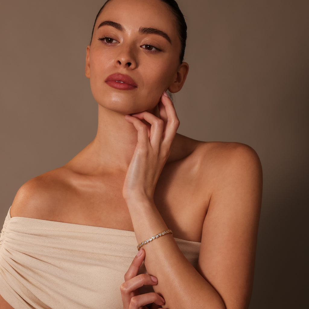A woman in an off-shoulder beige dress touches her neck, highlighting the Heart Tennis Bracelet in 18K Gold against a neutral brown background.
