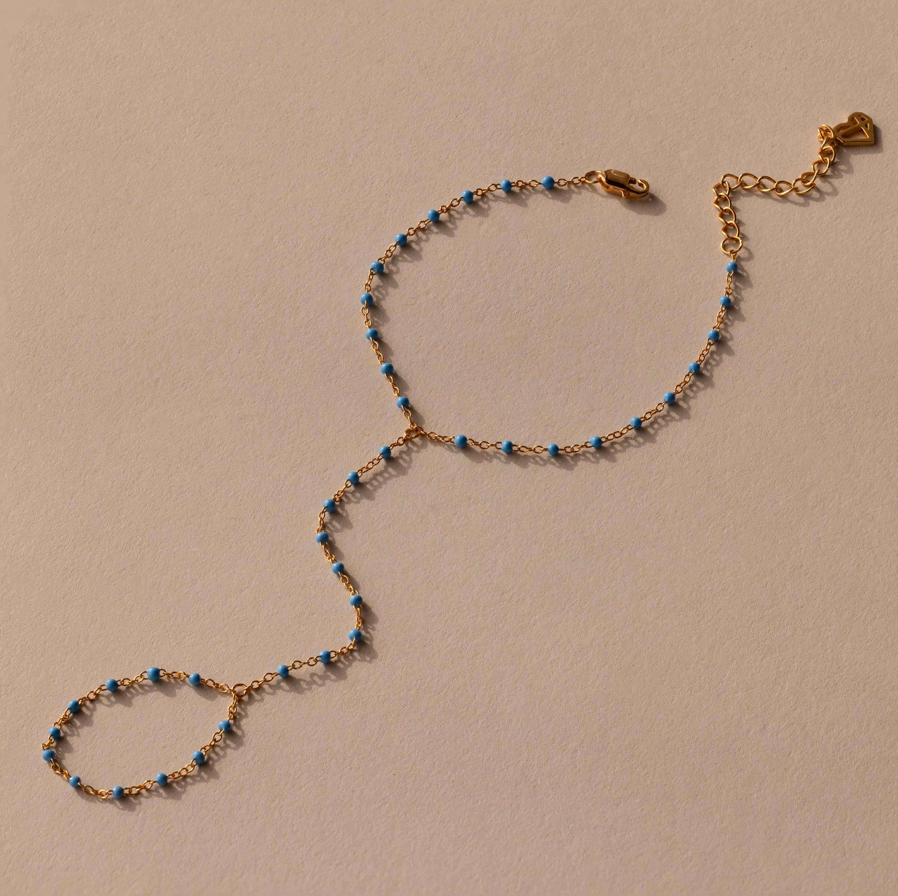 Ocean Beaded Hand Chain Bracelet in 18K Gold with blue beads, featuring a turquoise bracelet and ring linked by a delicate chain, showcased on a beige background.