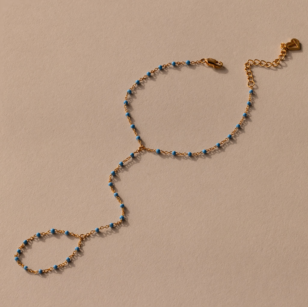 The Ocean Beaded Hand Chain Bracelet features a delicate gold chain with turquoise beads, an adjustable clasp, and ring detail, beautifully displayed on a neutral background.
