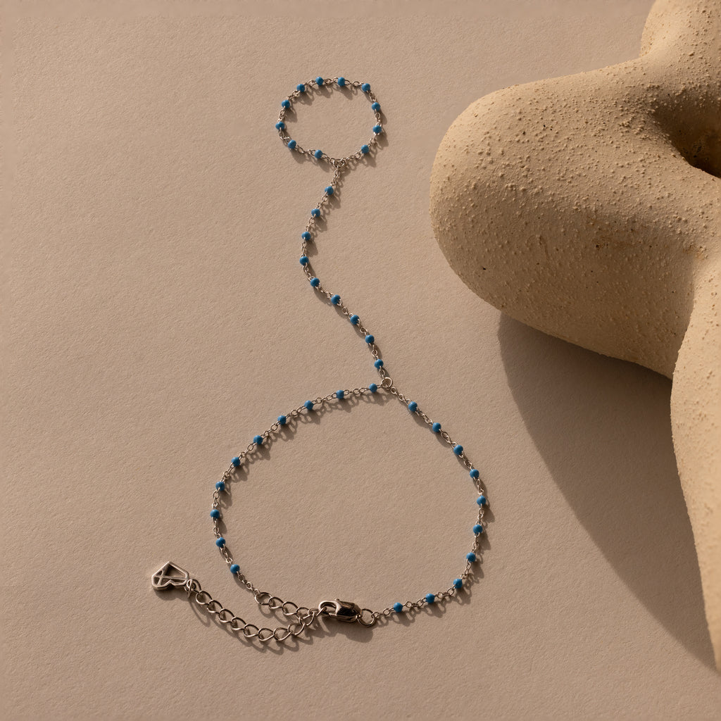 The Ocean Beaded Hand Chain Bracelet in Silver, featuring turquoise beads, is displayed on a beige textured surface next to a sculptural object.