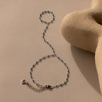 The Ocean Beaded Hand Chain Bracelet in Silver, featuring turquoise beads, is displayed on a beige textured surface next to a sculptural object.