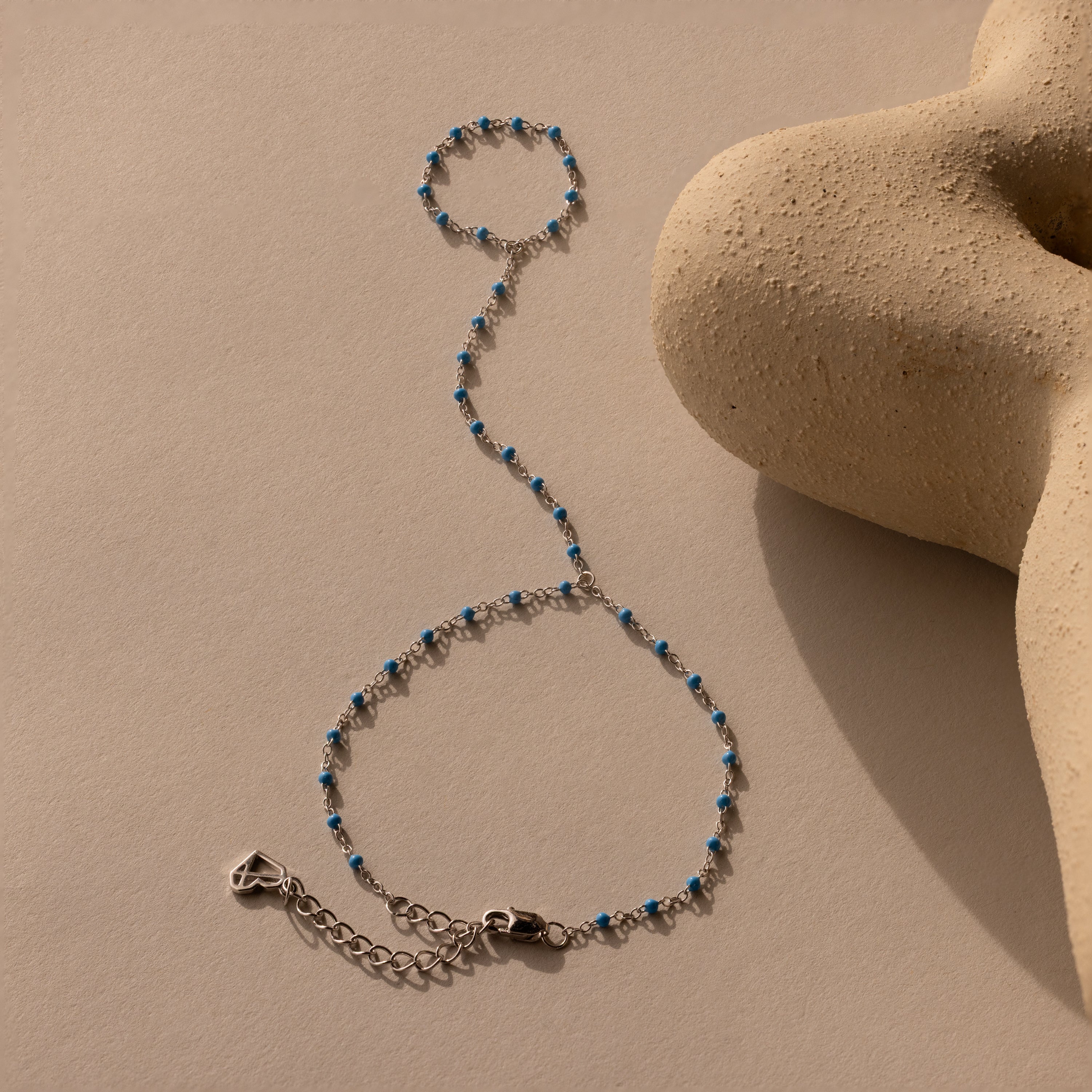 The Ocean Beaded Hand Chain Bracelet in Silver, featuring turquoise beads, is displayed on a beige textured surface next to a sculptural object.