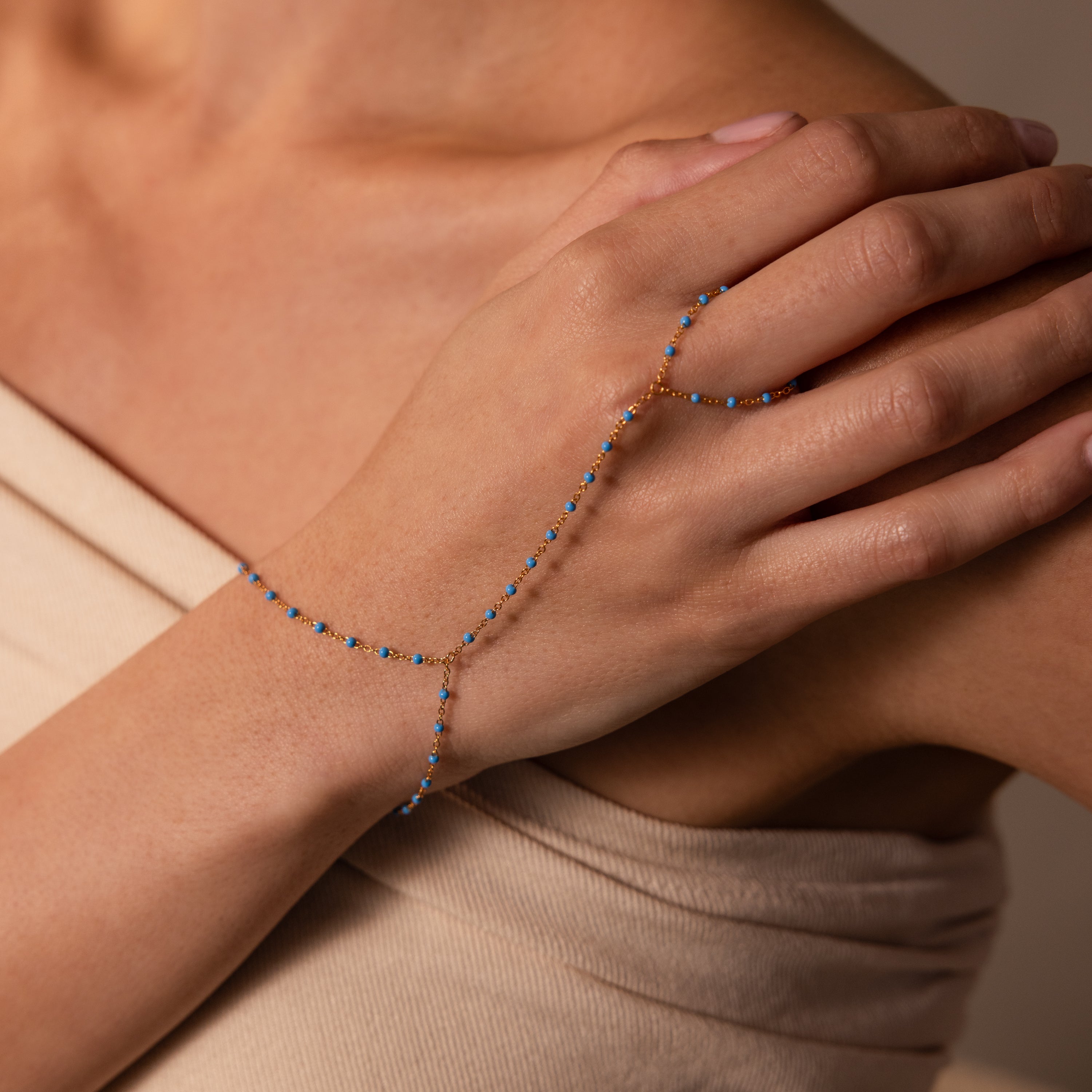 The Ocean Beaded Hand Chain Bracelet in 18K Gold features a delicate chain with blue beads that elegantly drapes across the hand and attaches to a ring, creating a refined, ocean-inspired accessory.