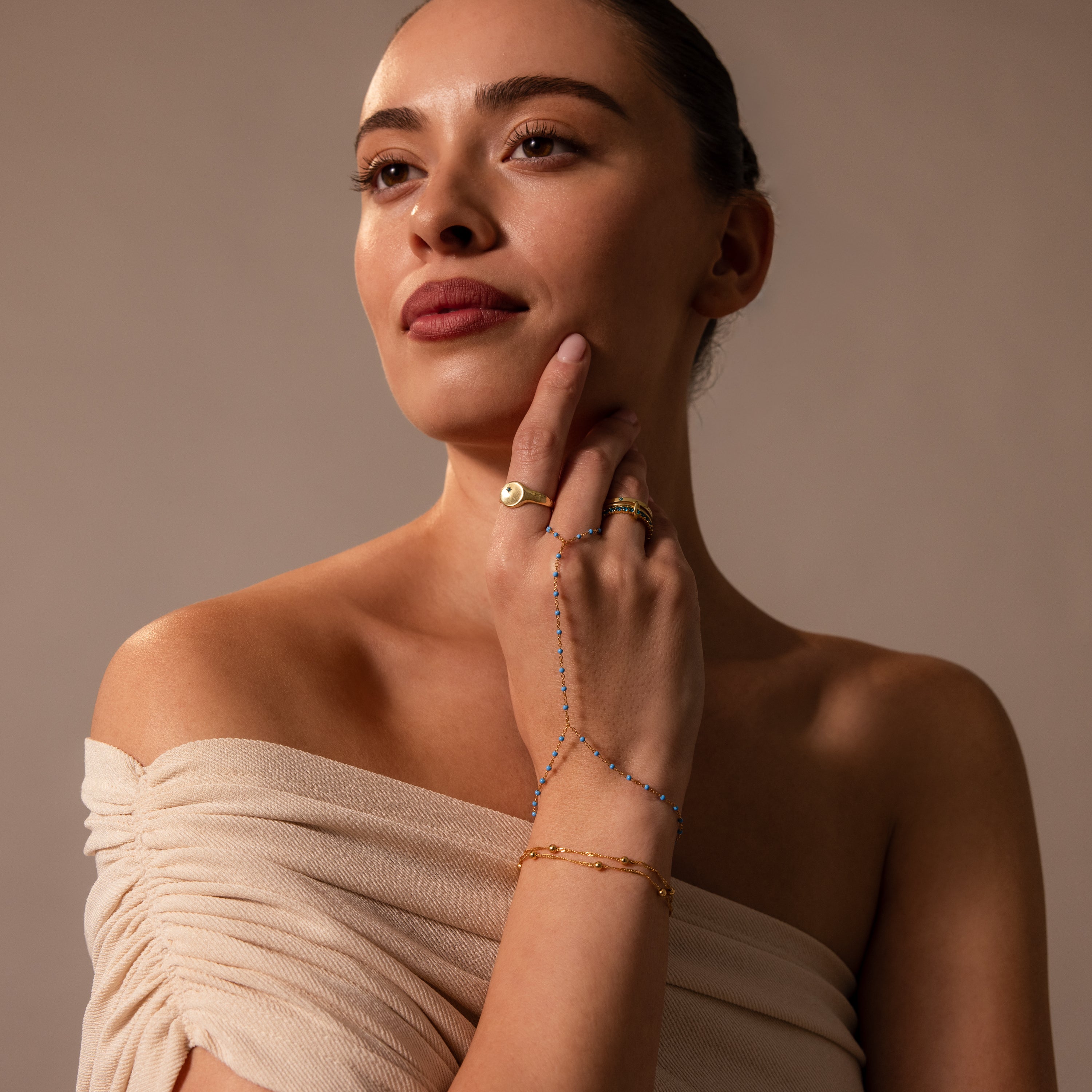 Woman in an off-shoulder beige top styled with gold rings, a turquoise bracelet, and the Ocean Beaded Hand Chain Bracelet in 18K Gold for a chic, layered look.