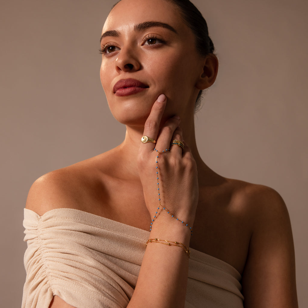 Woman in an off-the-shoulder beige top wears gold jewelry and the Ocean Beaded Hand Chain Bracelet, gazing thoughtfully to the side.