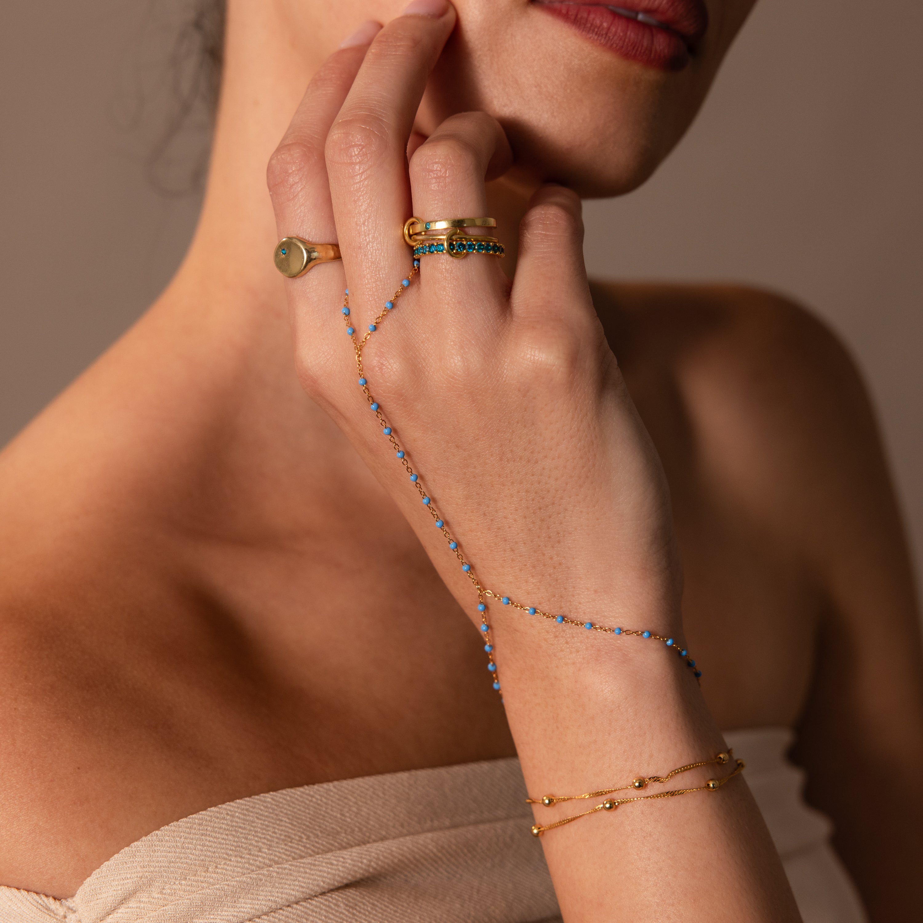 A woman wears gold rings and the Ocean Beaded Hand Chain Bracelet, its delicate blue beads adorning her hand as she touches her chin.