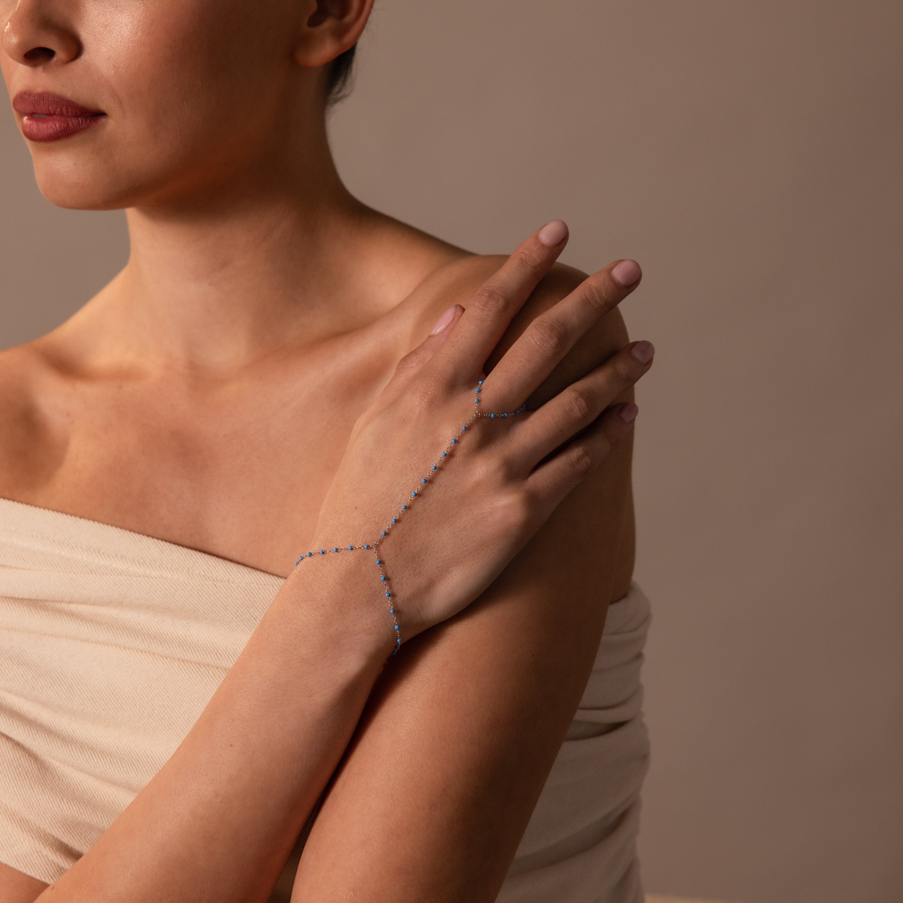 A woman in a beige top showcases the Ocean Beaded Hand Chain Bracelet in Silver, adorned with delicate turquoise beads and blue stones on her hand.