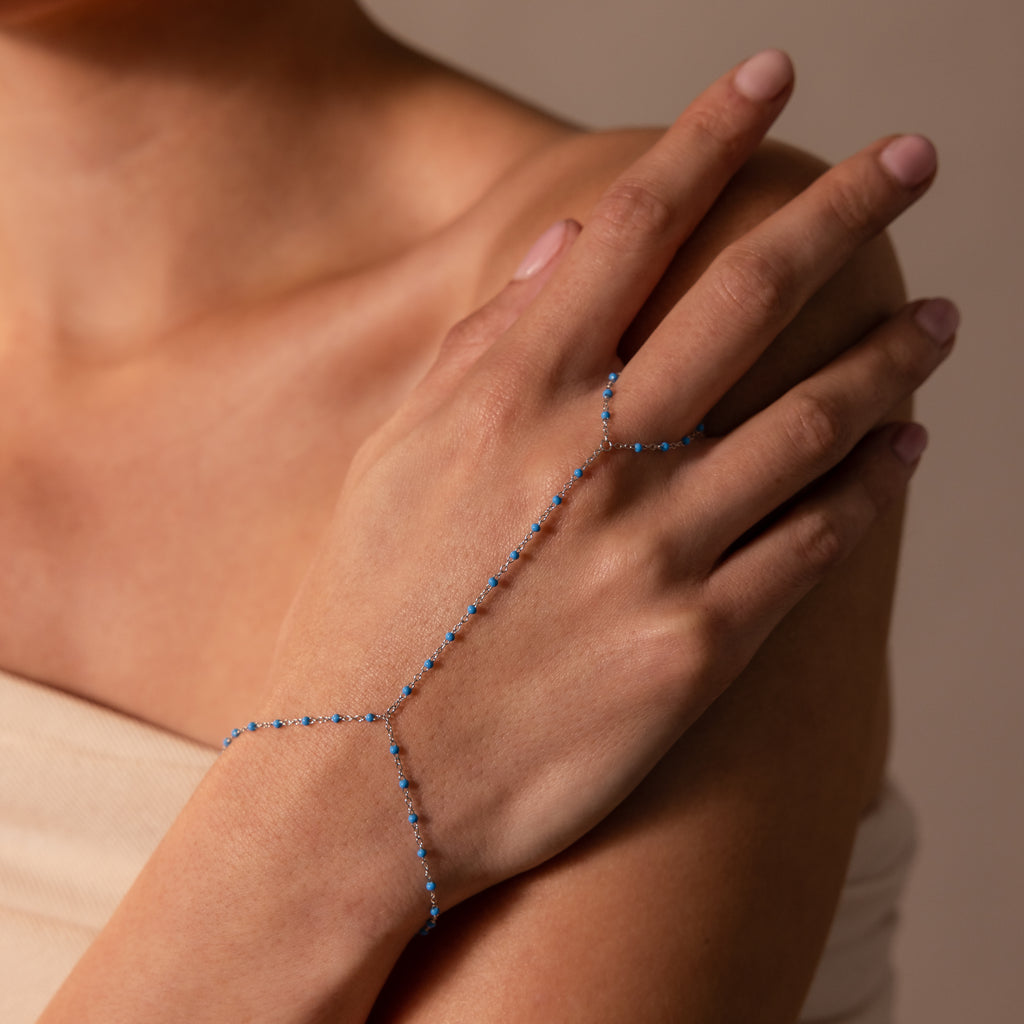 The Ocean Beaded Hand Chain Bracelet in Silver, featuring delicate turquoise beads, adorns a woman's hand and wrist set against a neutral background.