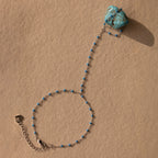 The Ocean Beaded Hand Chain Bracelet in Silver features blue and turquoise beads with a large turquoise stone, arranged on a textured beige surface for a touch of seaside elegance to any look.