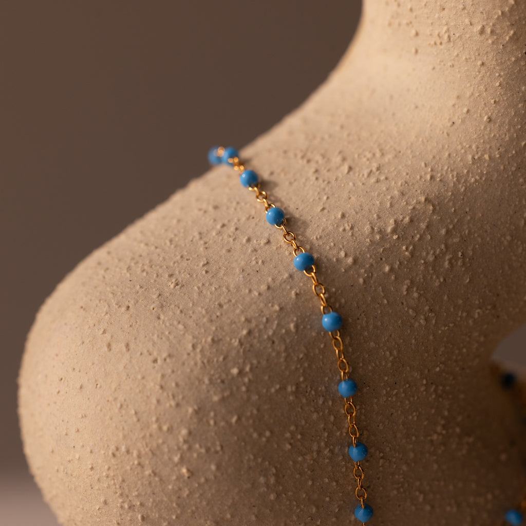 The Ocean Beaded Hand Chain Bracelet in 18K Gold features delicate chains with small blue beads, draped elegantly over a textured beige sculpture shaped like a human shoulder, echoing ocean-inspired sophistication.