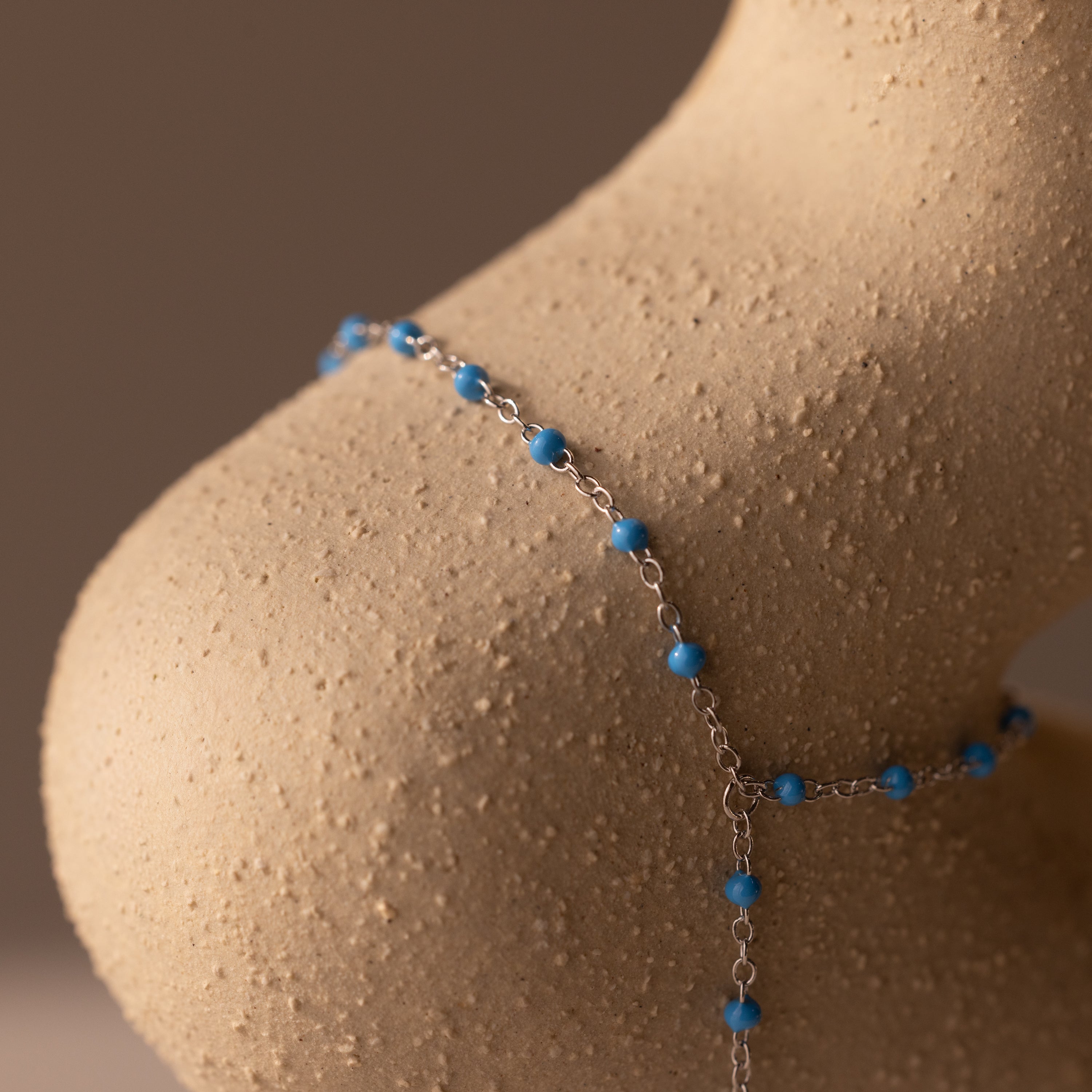 The Ocean Beaded Hand Chain Bracelet in Silver, adorned with turquoise beads, drapes elegantly over a textured beige sculpture shaped like a human shoulder.