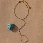 The Ocean Beaded Hand Chain Bracelet in 18K Gold, adorned with small blue beads, is displayed next to a turquoise stone on a beige textured surface.