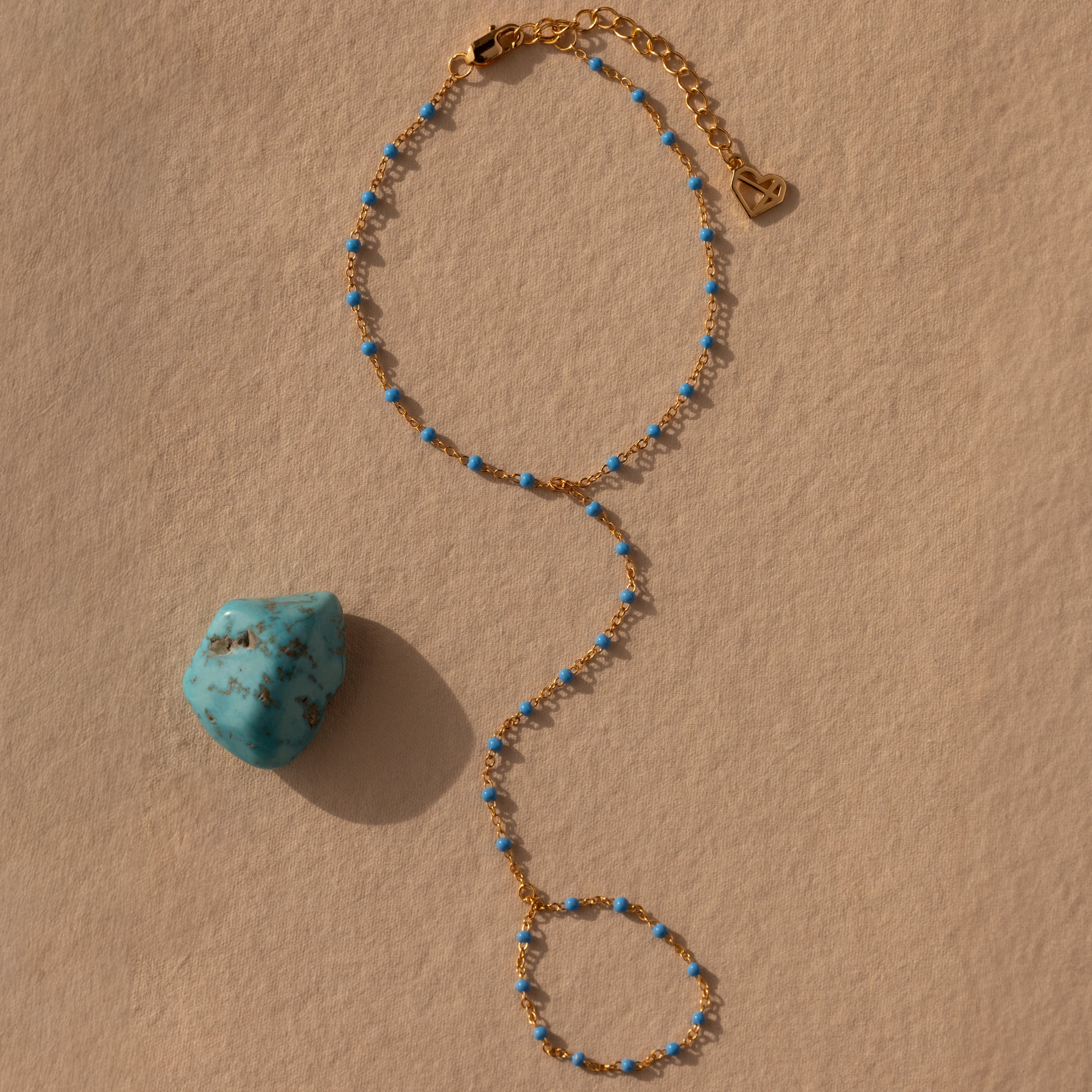 The Ocean Beaded Hand Chain Bracelet, a gold delicate chain with small blue beads, rests by turquoise on a beige textured surface, capturing the charm of ocean-inspired jewelry.