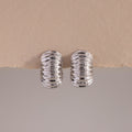A pair of Chunky Ribbed Hoops in white gold, positioned upright and symmetrically on a lightly textured, torn-edge beige surface. The hoops have a bold, rounded silhouette with a series of prominent, high-polish ridges that create a corrugated effect