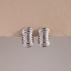 A pair of Chunky Ribbed Hoops in white gold, positioned upright and symmetrically on a lightly textured, torn-edge beige surface. The hoops have a bold, rounded silhouette with a series of prominent, high-polish ridges that create a corrugated effect