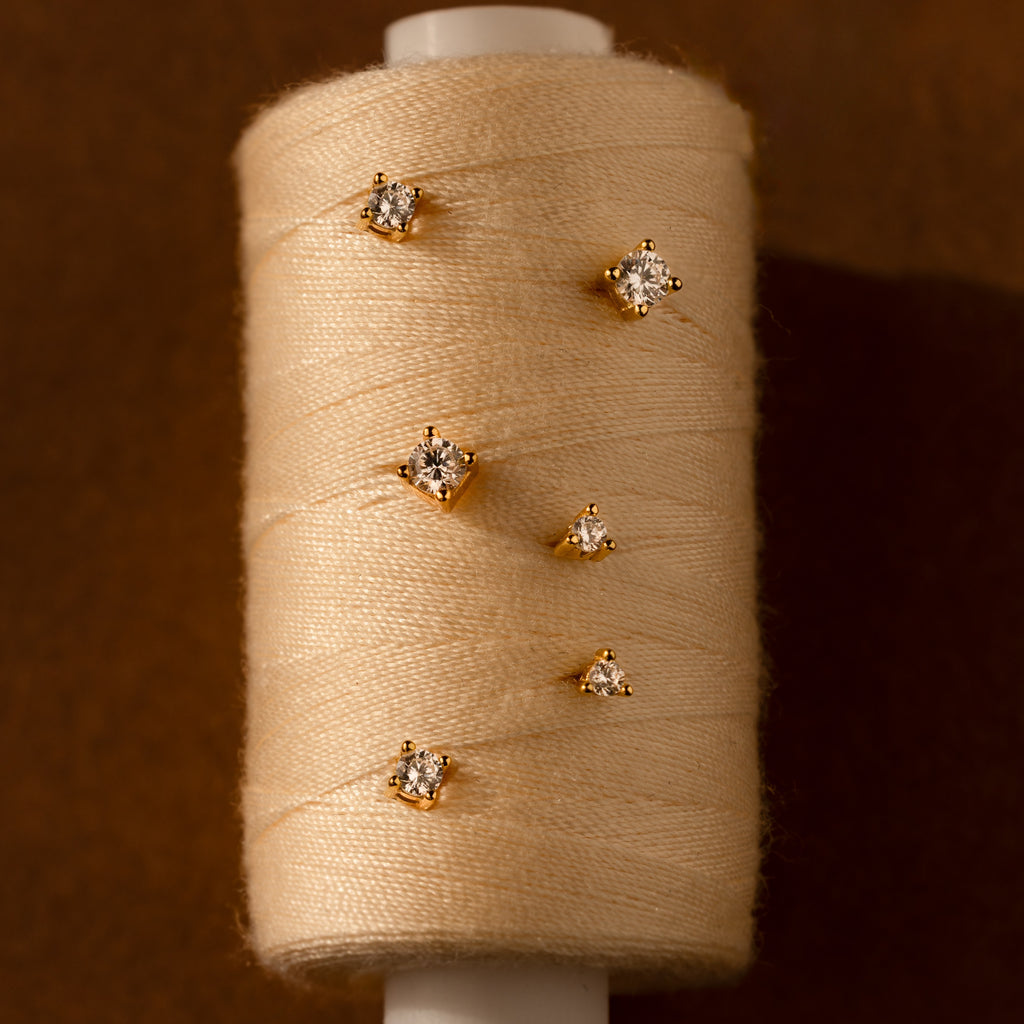 The Booker Large Diamond Studs—five round-cut gold diamonds—shine against beige thread and a brown background, making them the perfect everyday earrings.
