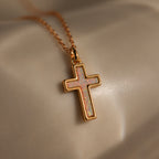The Mother of Pearl Cross Necklace in 18K Gold, featuring an iridescent inlay pendant, rests on a smooth beige surface—a timeless jewelry piece perfect for any occasion.