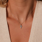 The Mother of Pearl Cross Necklace in Silver rests on her neck, its iridescent inlay glowing softly against a beige knit top with a deep neckline—a timeless piece of jewelry.