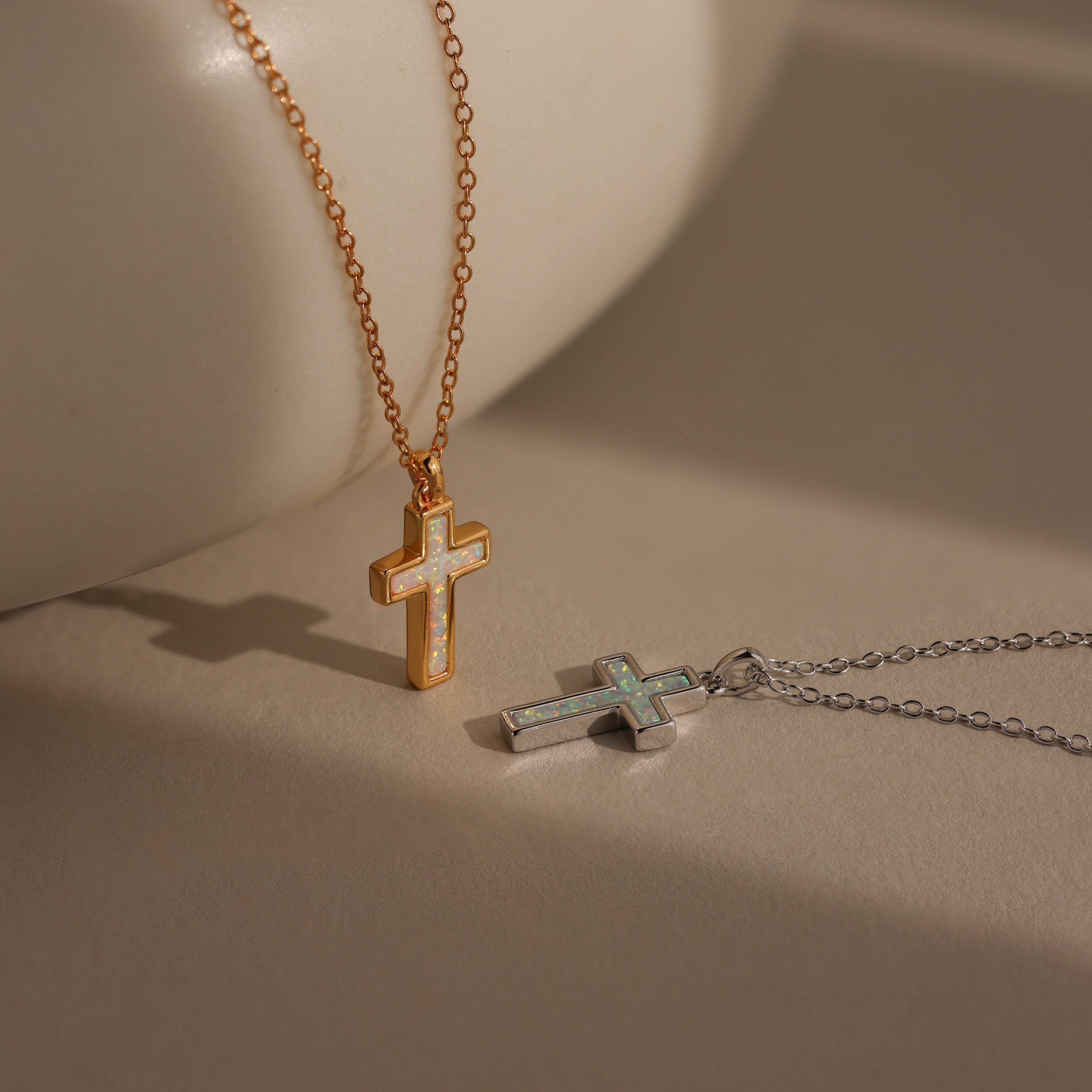 The Mother of Pearl Cross Necklace is shown in both gold and silver on a neutral surface with soft shadows. One necklace features an iridescent inlay that catches the light, making it a timeless piece for any occasion.