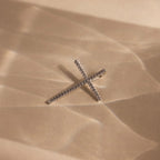 The Skinny Pave Cross Charm in Silver, sparkling with diamonds, lies on textured beige sand highlighted by soft shadows.