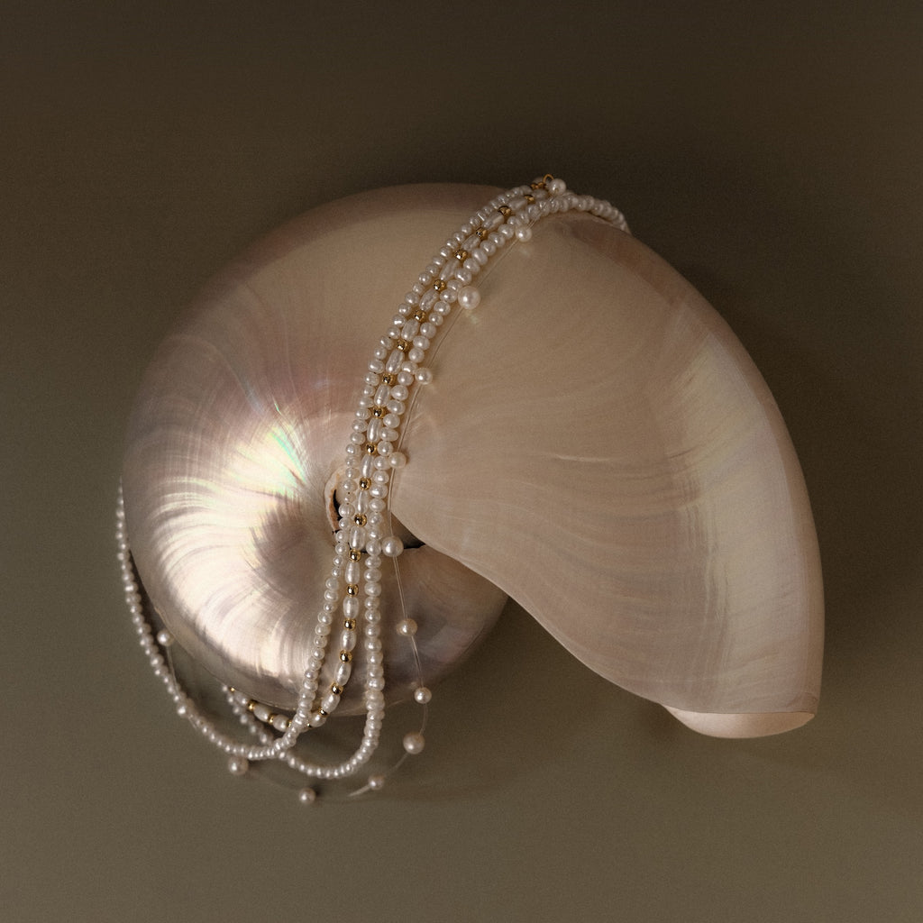 The Summer Pearl Beaded Necklace in 18K Gold is draped elegantly over a shiny spiral seashell on a simple neutral background.