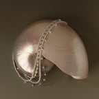 The Summer Pearl Beaded Necklace in 18K Gold is draped elegantly over a shiny spiral seashell on a simple neutral background.