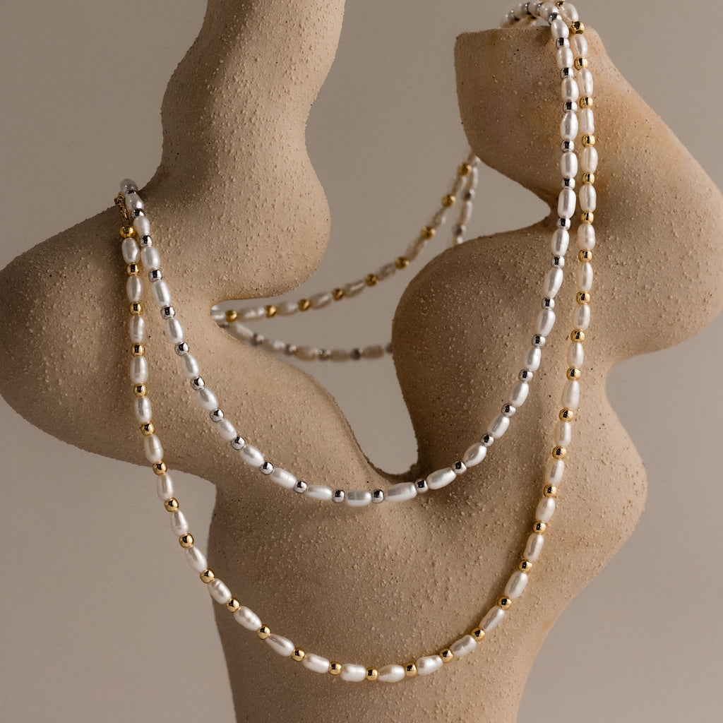 Two strands of polished beads and gold drape elegantly over an abstract beige sculpture, showcasing the Beaded Pearl Necklace with graceful freshwater pearls.