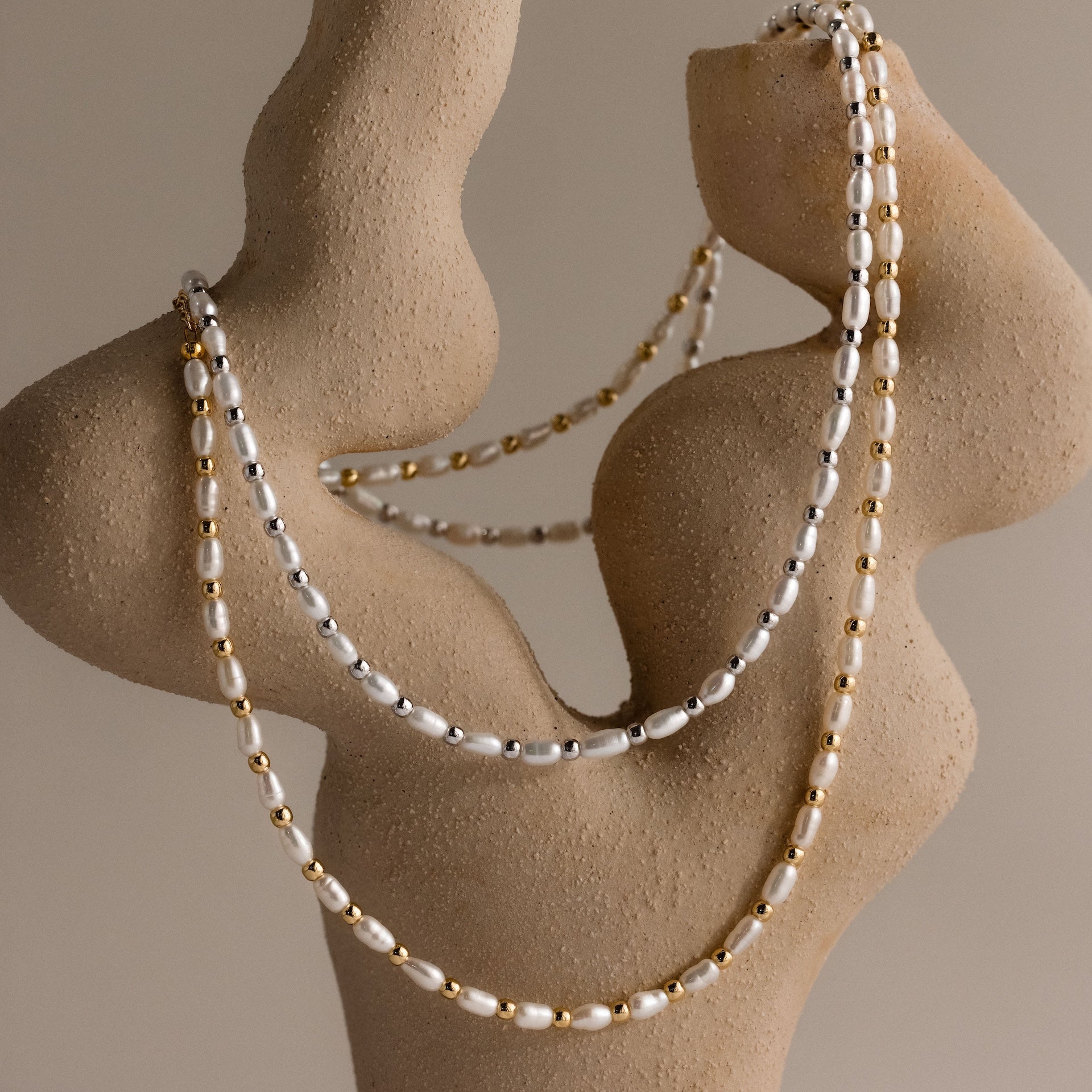 Two strands of polished beads and gold drape elegantly over an abstract beige sculpture, showcasing the Beaded Pearl Necklace with graceful freshwater pearls.