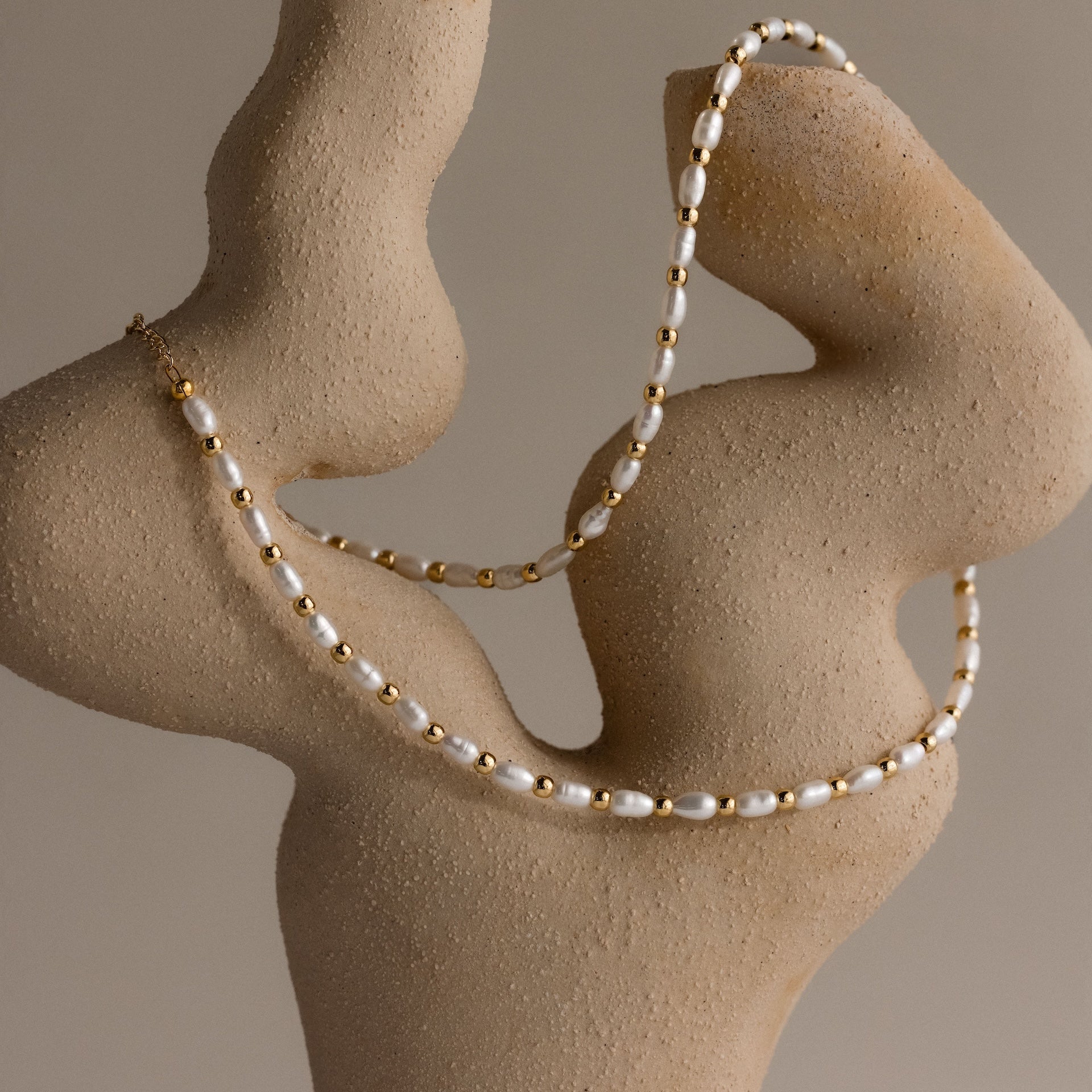 The Beaded Pearl Necklace in 18K Gold, featuring delicate polished beads and freshwater pearls, is elegantly draped over a textured, abstract beige sculpture.