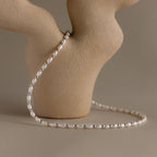 The Beaded Pearl Necklace, featuring delicate polished beads, is draped over a textured beige ceramic vase with a neutral background.