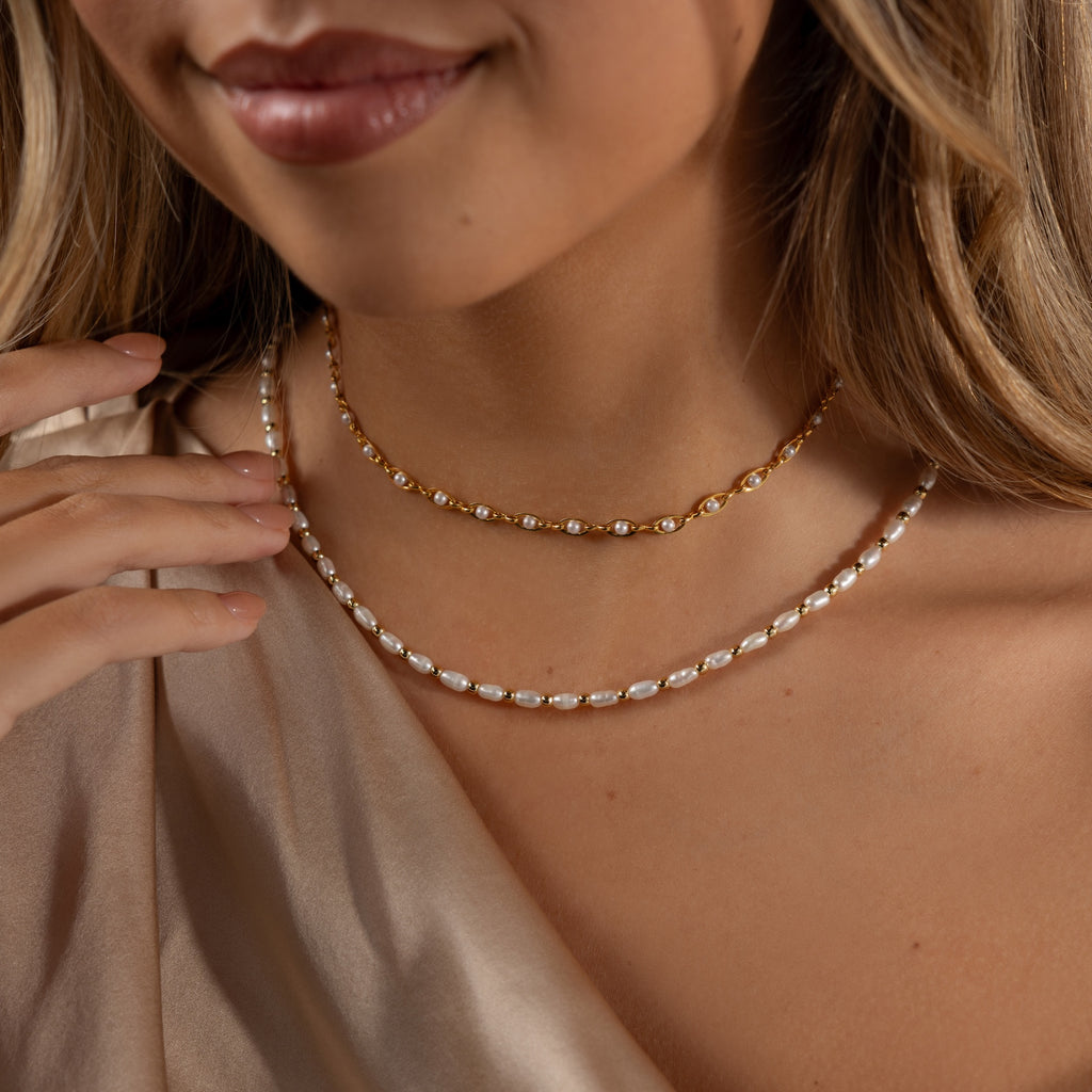 A woman smiles in a close-up shot, wearing the Beaded Pearl Necklace in 18K Gold layered over a cream off-the-shoulder satin top.
