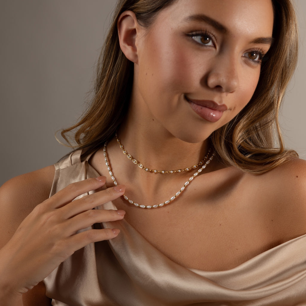 A woman in a satin dress wears layered gold and pearl necklaces, including the Beaded Pearl Necklace in 18K Gold with delicate polished beads and freshwater pearls, gently touching one of the necklaces.